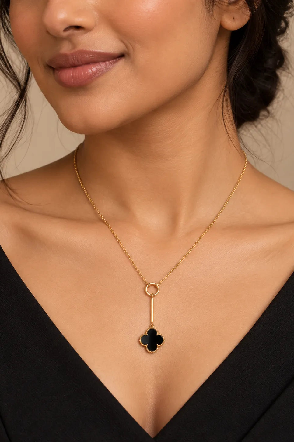 A close-up of a woman wearing a delicate gold necklace with a black clover pendant, styled with a black V-neck top against a warm beige background.