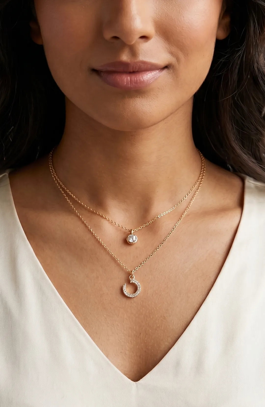 A close-up of a woman wearing two delicate gold necklaces: one with a small round clear stone pendant and the other with a small halo-shaped pendant, paired with a white V-neck top.