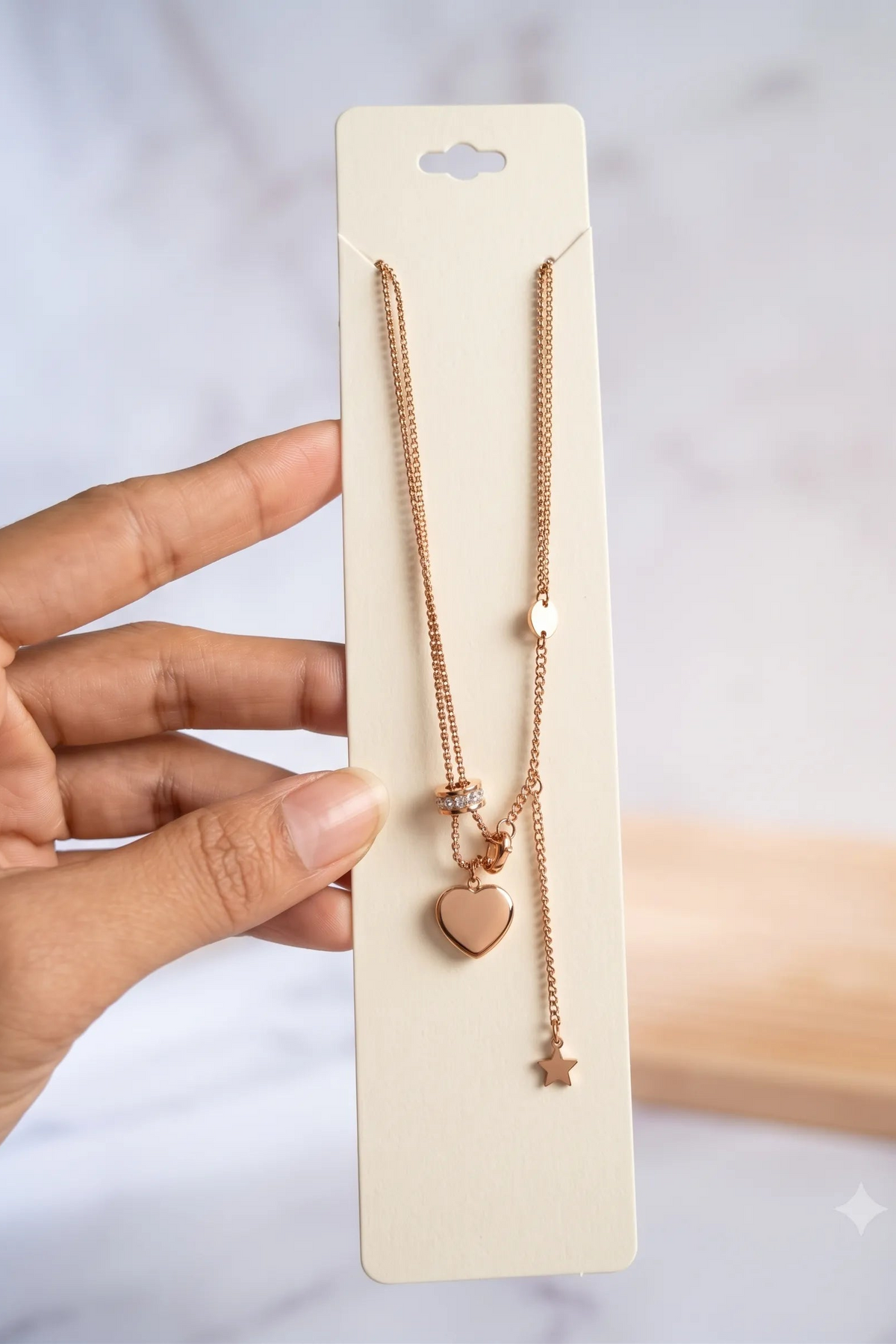 A rose gold double-strand necklace with a heart and star pendant, displayed on a cream-colored card and held by a hand against a soft marble background.