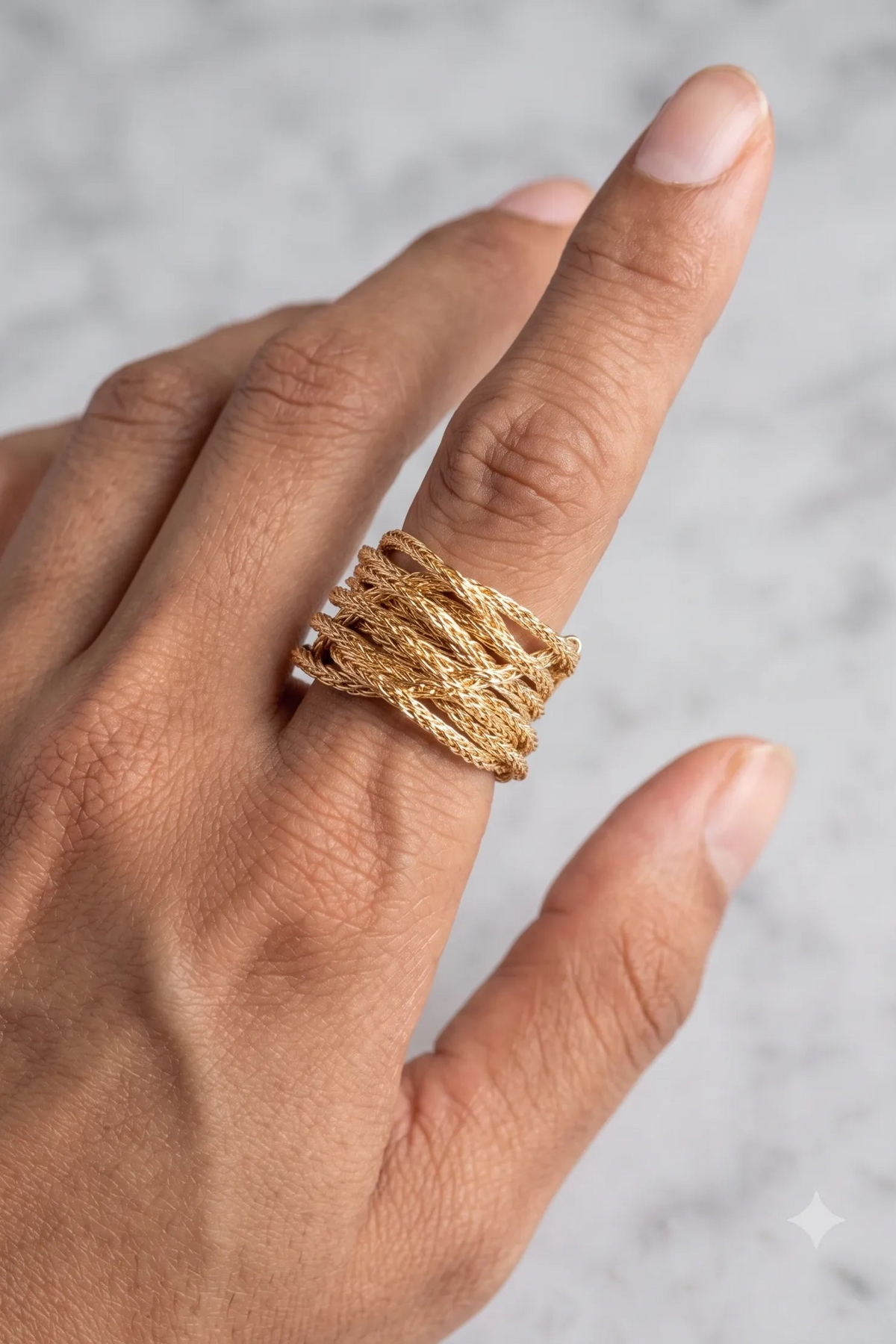 A close-up of a hand wearing a wide, multi-strand textured gold ring against a light gray marble background.