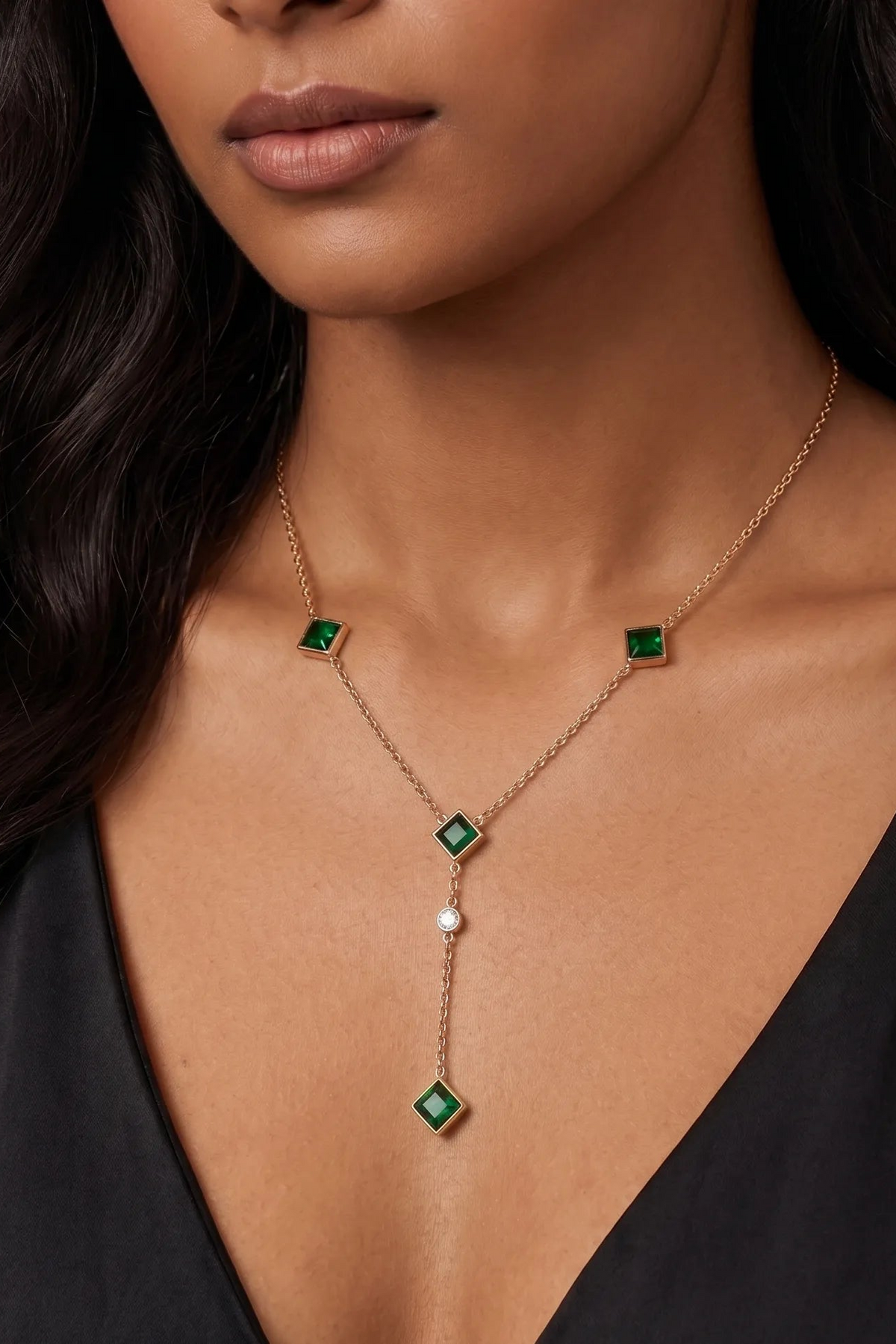 A close-up of a model wearing a delicate gold Y-necklace with three green diamond-shaped emerald-cut stones and a small white pearl accent.