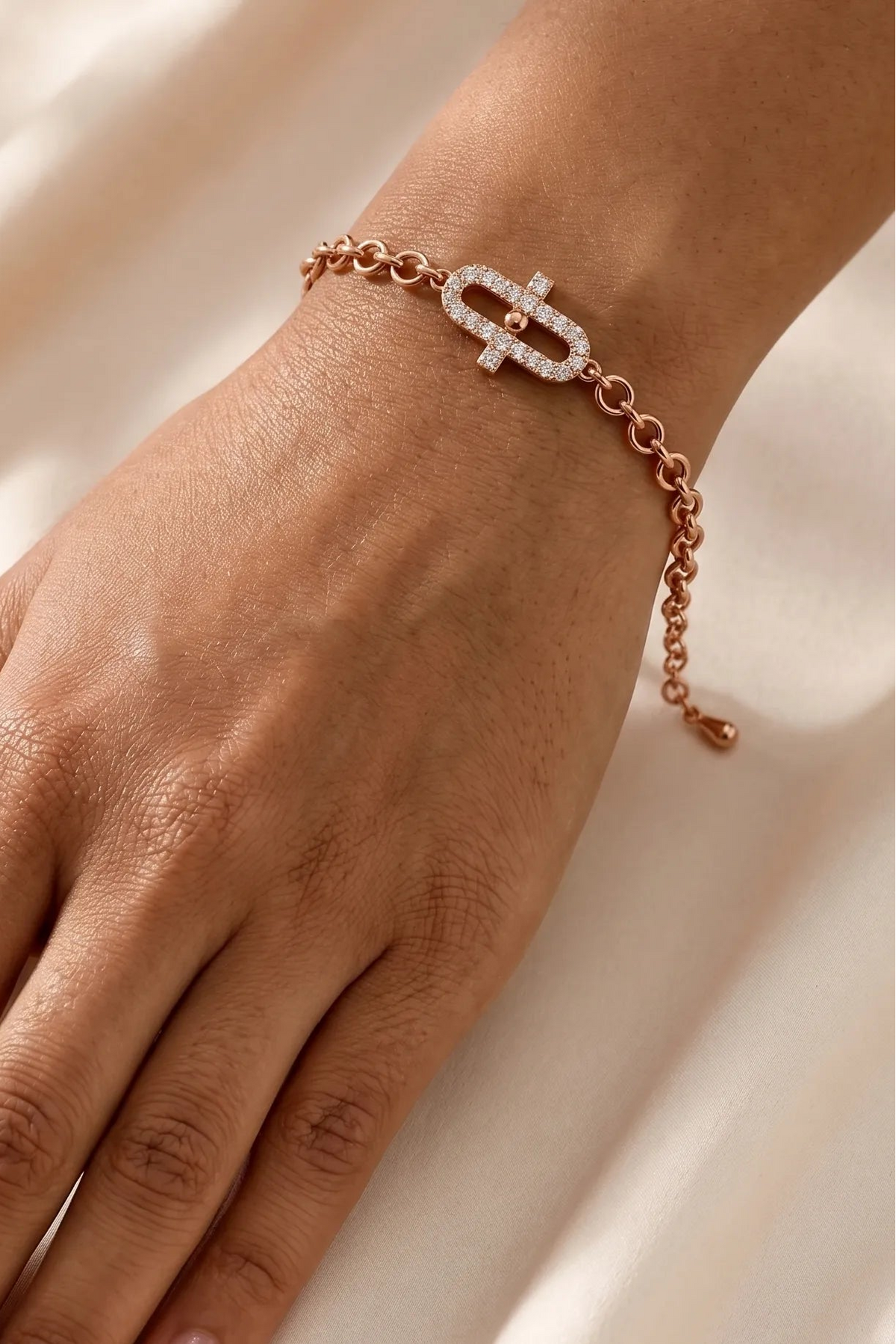 A close-up of a person's hand wearing a delicate rose gold chain bracelet with a geometric link design set with clear stones, resting on a soft, draped cream fabric background.