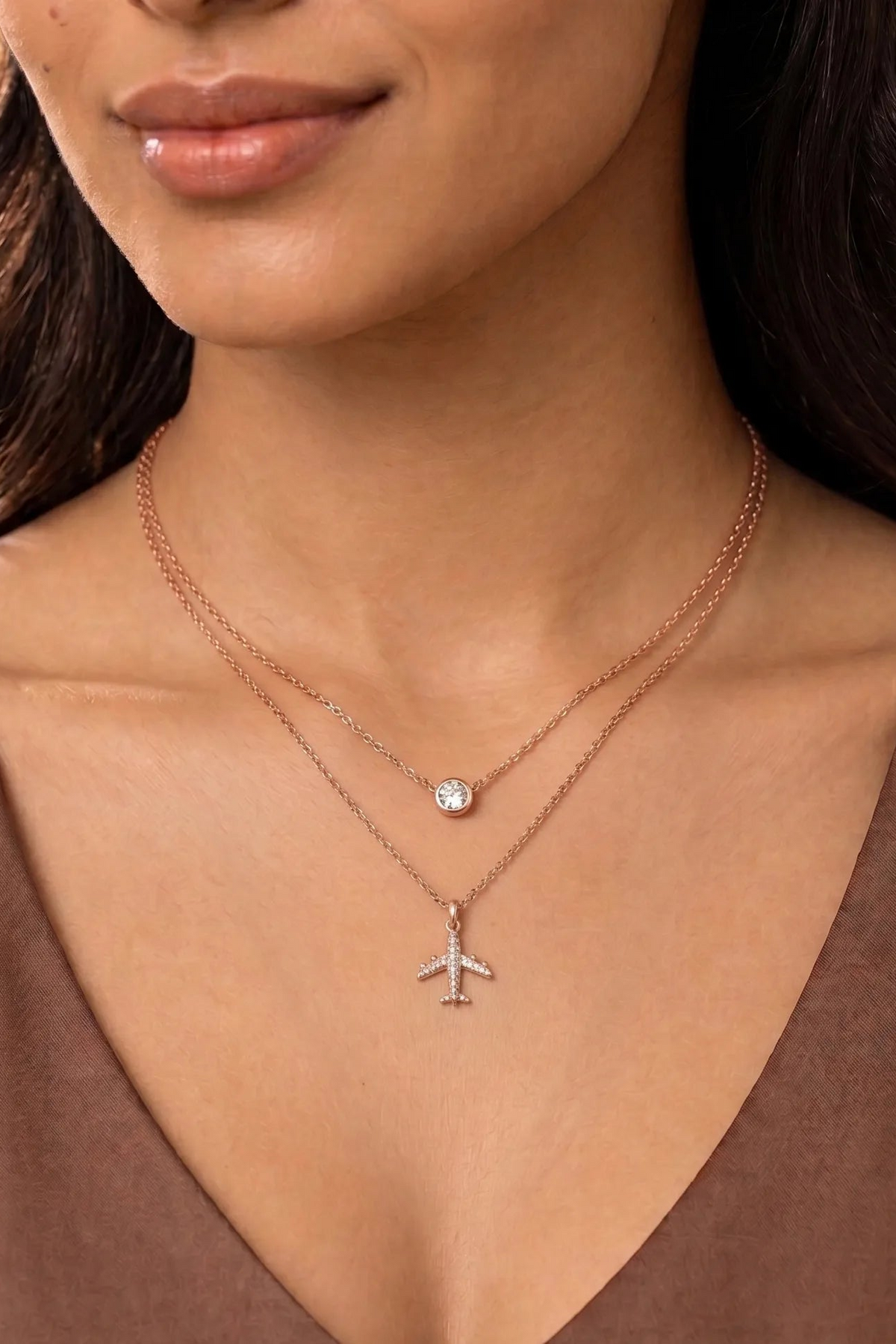 A close-up of a model wearing a double-layered rose gold necklace. The top layer features a small round bezel-set clear gemstone, and the bottom layer features a delicate airplane pendant encrusted with small clear stones.