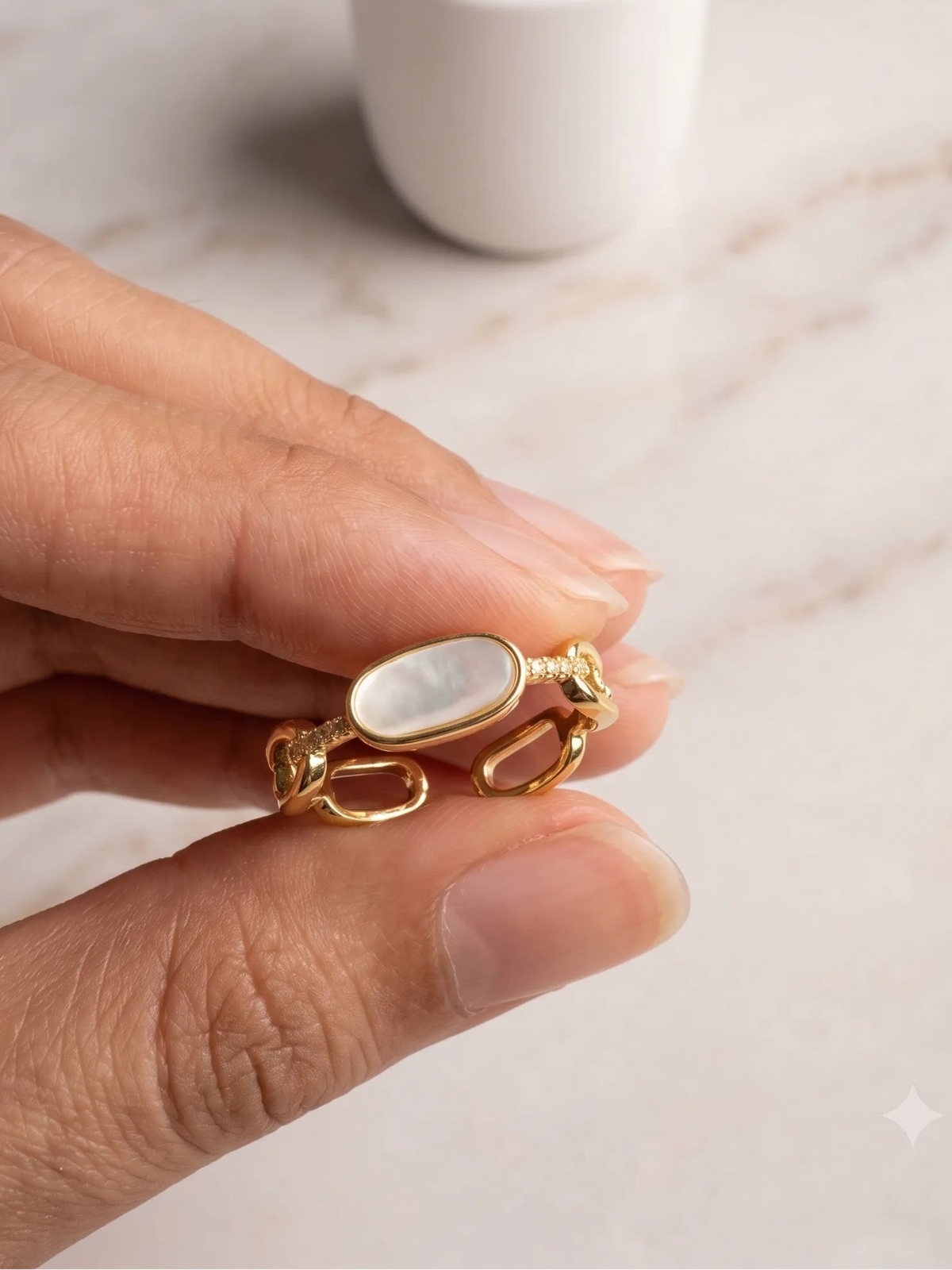 A hand holding a gold ring with an oval white mother-of-pearl inlay, with a white ceramic candle in the blurred background on a light marble surface.