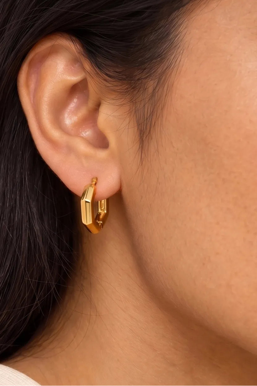A close-up of a model's ear wearing a small gold hoop earring with a geometric, faceted rectangular design.