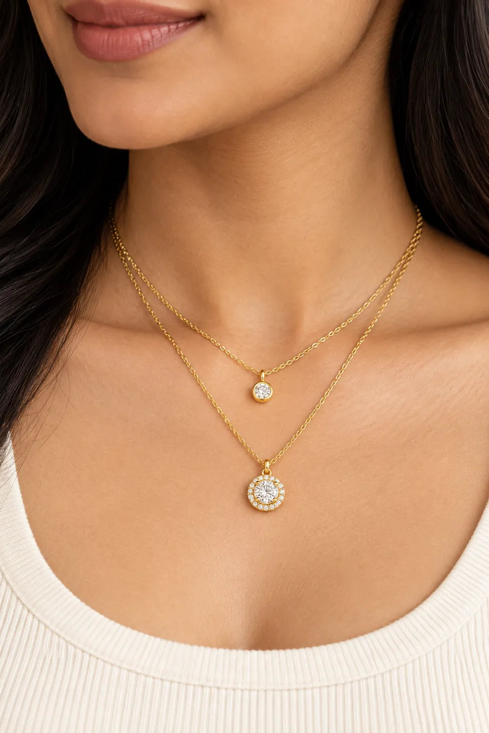 A close-up of a model wearing a double-layered gold necklace set. The set features a delicate chain with a small round bezel-set clear stone pendant and a thicker chain with a larger round halo-style pendant containing clear stones.