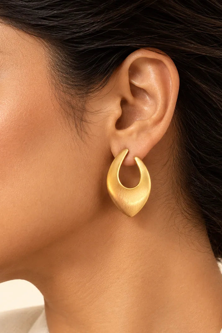 A close-up side profile of a woman wearing a single matte gold crescent-shaped hoop earring against a soft cream background.