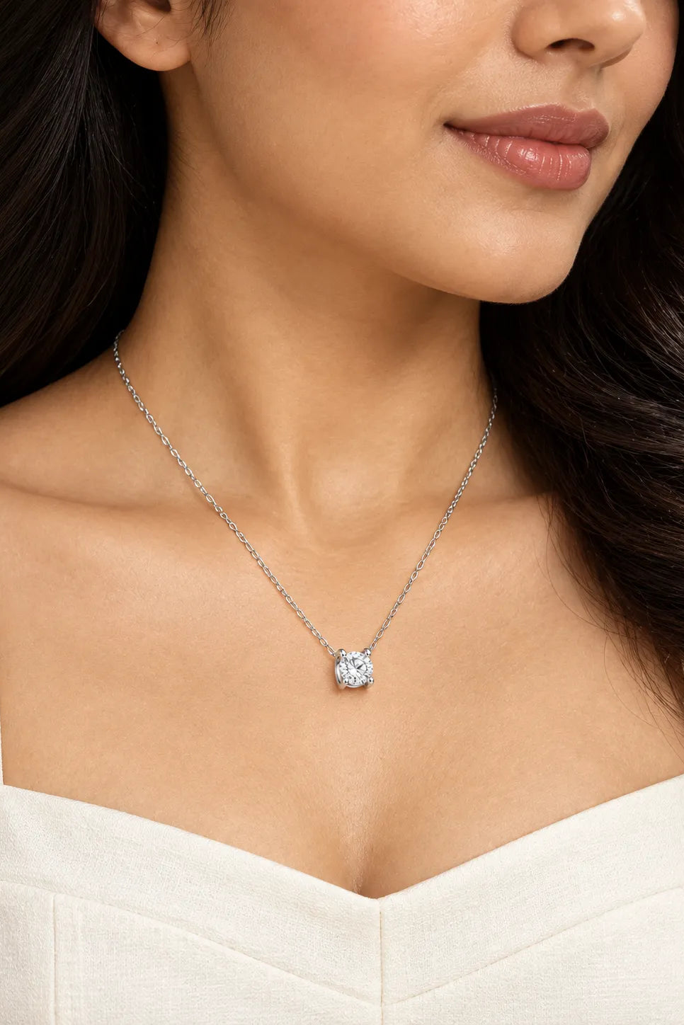 A close-up of a woman wearing a delicate silver chain necklace with a single round-cut clear gemstone pendant, styled with a white top.