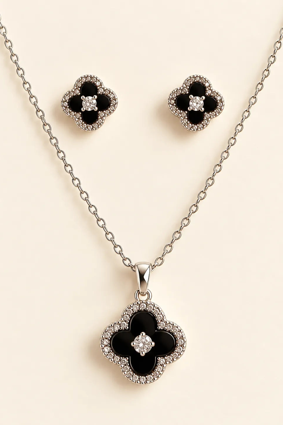 Silver-tone jewelry set featuring a necklace and matching stud earrings with black enamel clover motifs surrounded by clear rhinestones, displayed on a light beige background.