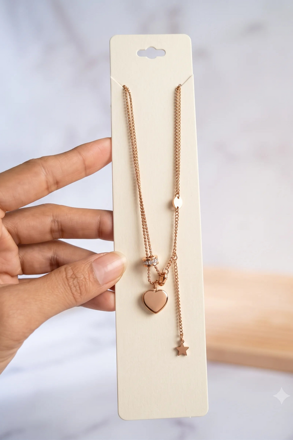 A rose gold double-strand necklace with a heart and star pendant, displayed on a cream-colored card and held by a hand against a soft marble background.