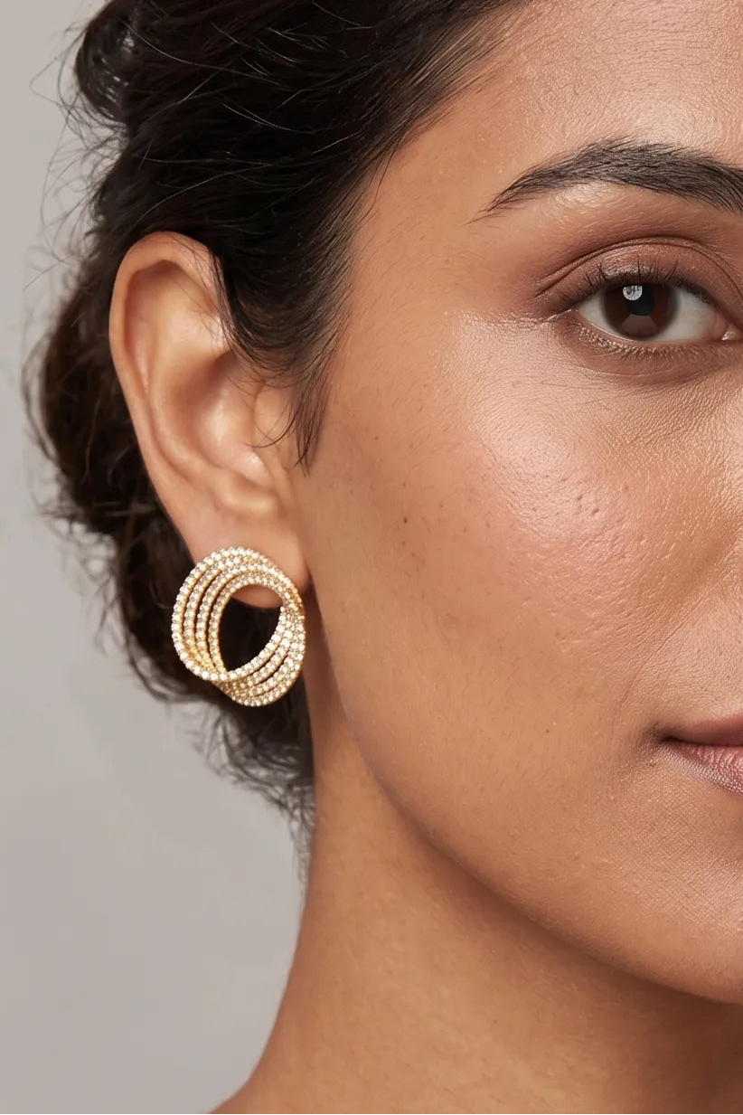 A close-up of a woman's ear wearing a gold-toned triple-ring hoop earring with textured, pave-set details, set against a neutral gray background.