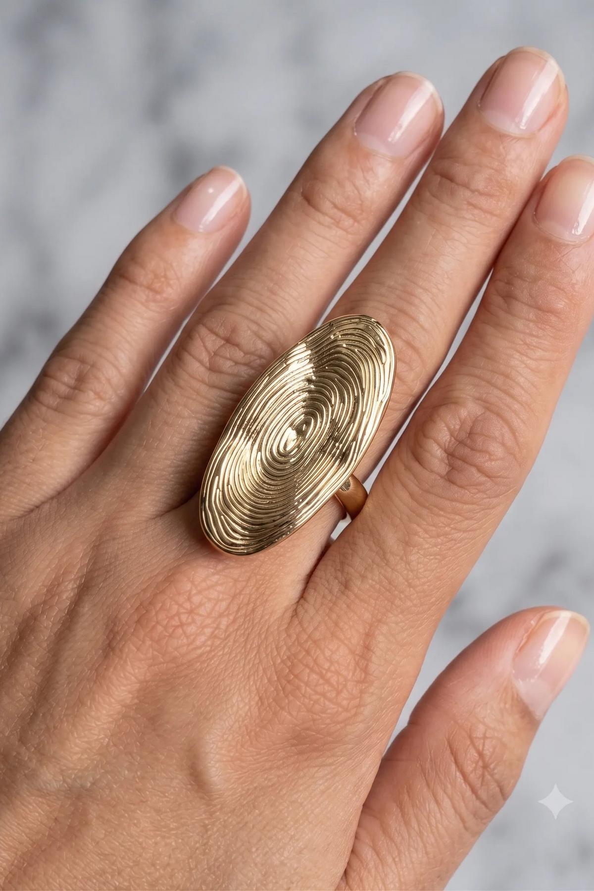 A close-up of a hand wearing a large oval gold ring with concentric spiral detailing, set against a light gray marble background.