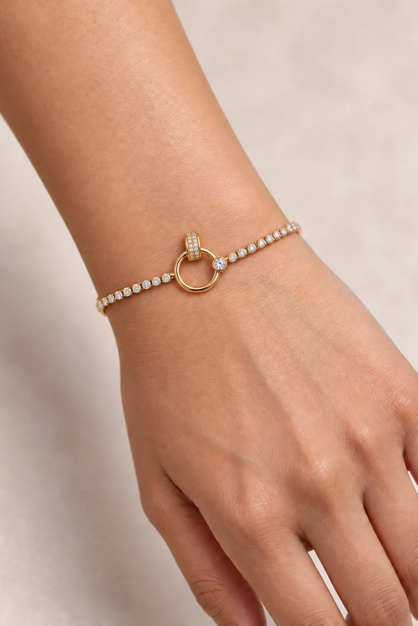 A delicate gold bracelet with a central gold ring and small clear stones, shown worn on a person's wrist against a soft beige background.