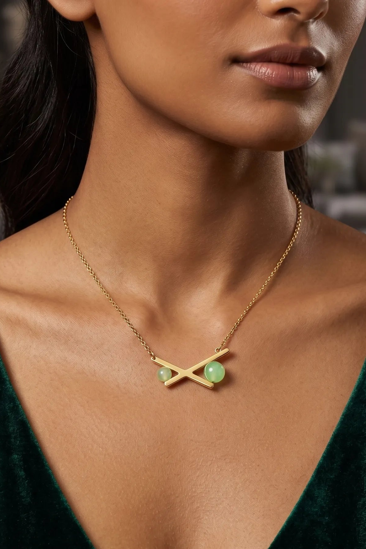 A close-up of a model wearing a delicate gold chain necklace with a geometric cross pendant featuring two light green oval stones, paired with a dark green velvet top.