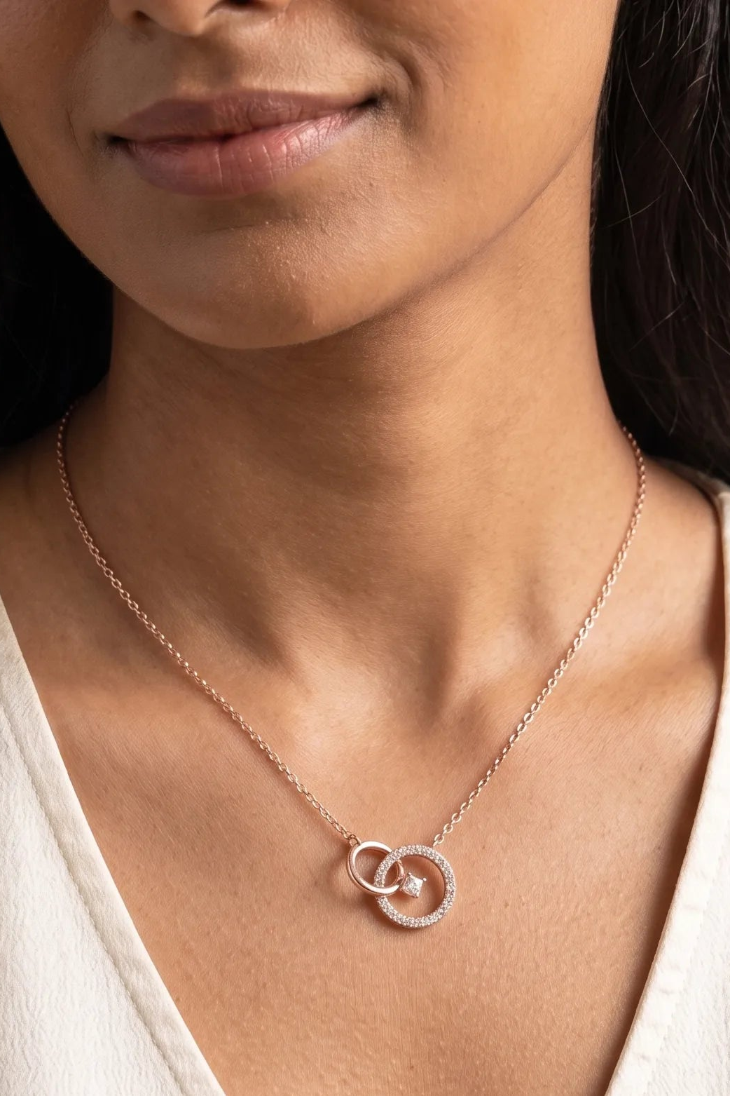 A close-up of a woman wearing a delicate rose gold chain necklace with interlocking double rings, one of which is adorned with small clear stones.