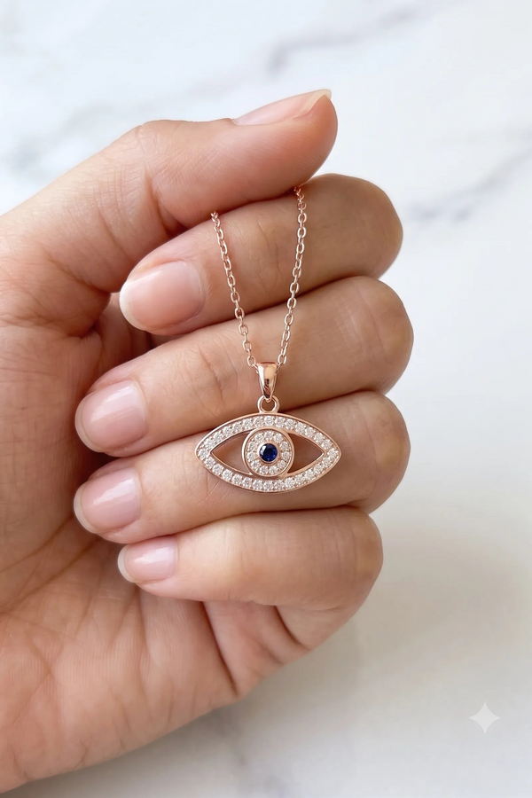 A hand holding a rose gold evil eye pendant necklace with a blue center stone and clear stones around it, set against a light marble background.