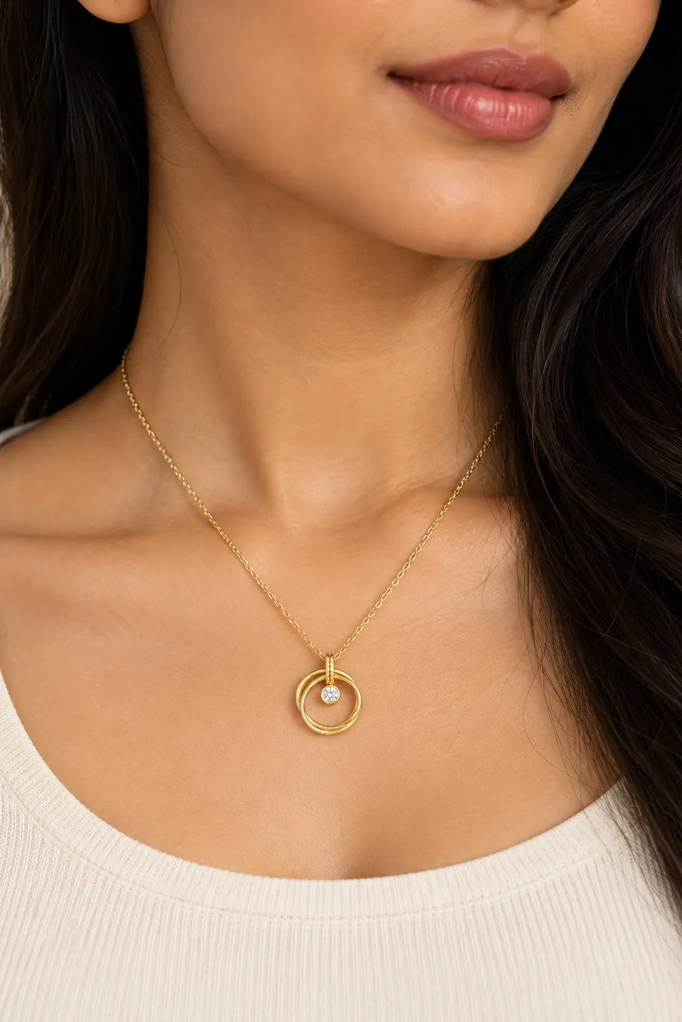 A close-up of a woman wearing a delicate gold necklace with a double-ring pendant and a small clear stone, paired with a ribbed cream top.