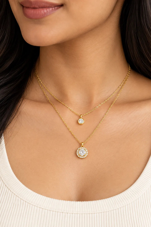 A close-up of a model wearing a double-layered gold necklace set. The set features a delicate chain with a small round bezel-set clear stone pendant and a thicker chain with a larger round halo-style pendant containing clear stones.