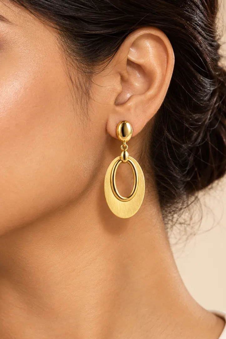 A close-up of a woman wearing a gold drop earring with an oval design, shown against a warm beige background.