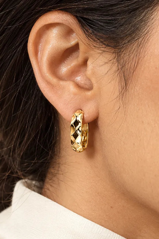 A close-up of a model's ear wearing a small gold huggie hoop earring with a textured, faceted geometric pattern.