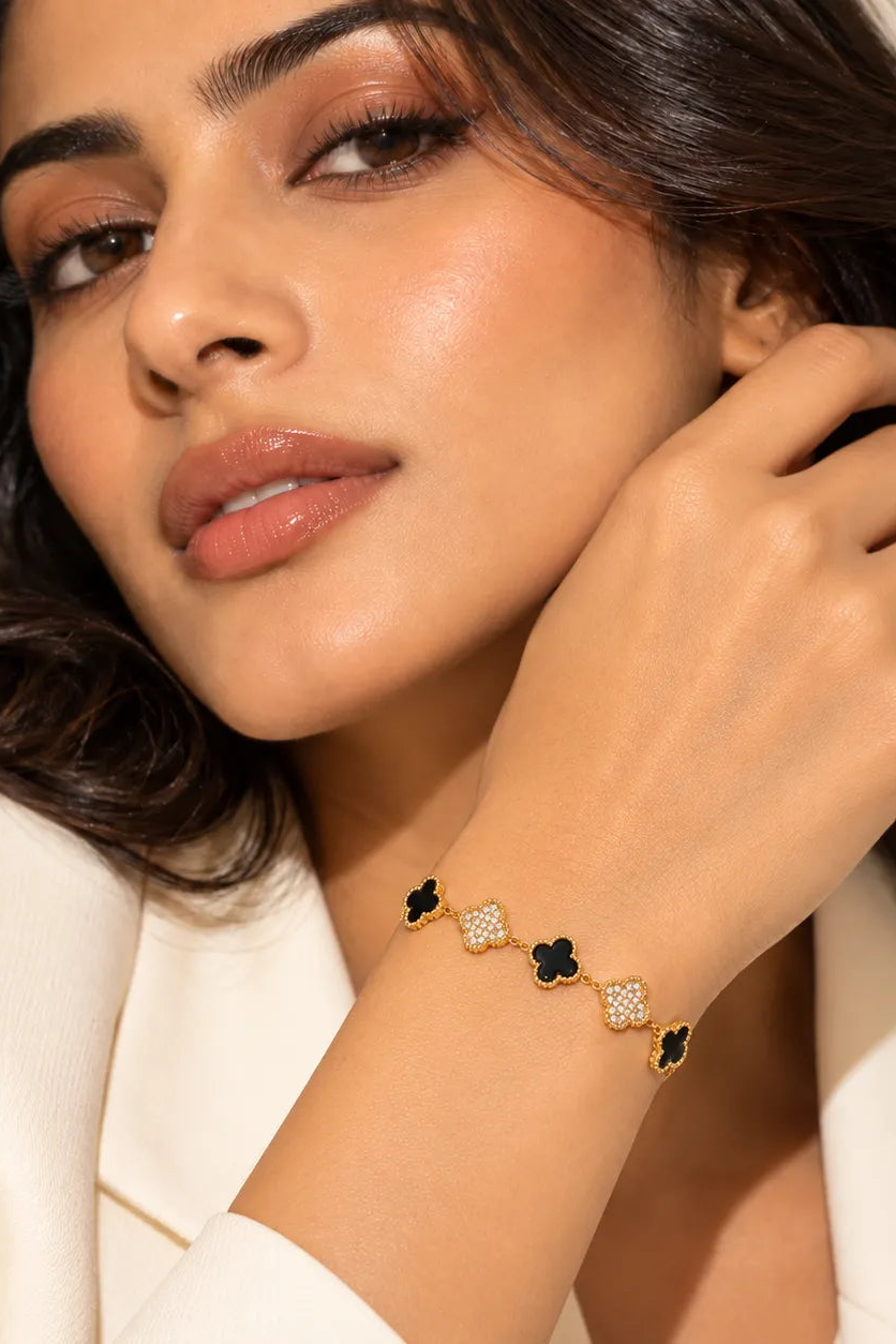 A close-up of a woman wearing a delicate bracelet with alternating black clover and crystal-encrusted gold clovers, paired with a cream-colored blazer.