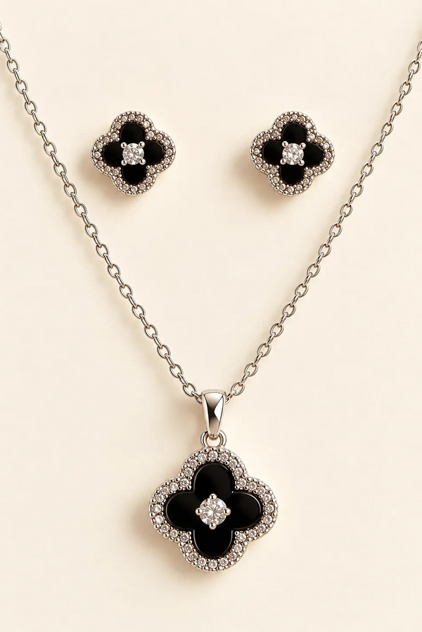 Silver-tone jewelry set featuring a necklace and matching stud earrings with black enamel clover motifs surrounded by clear rhinestones, displayed on a light beige background.