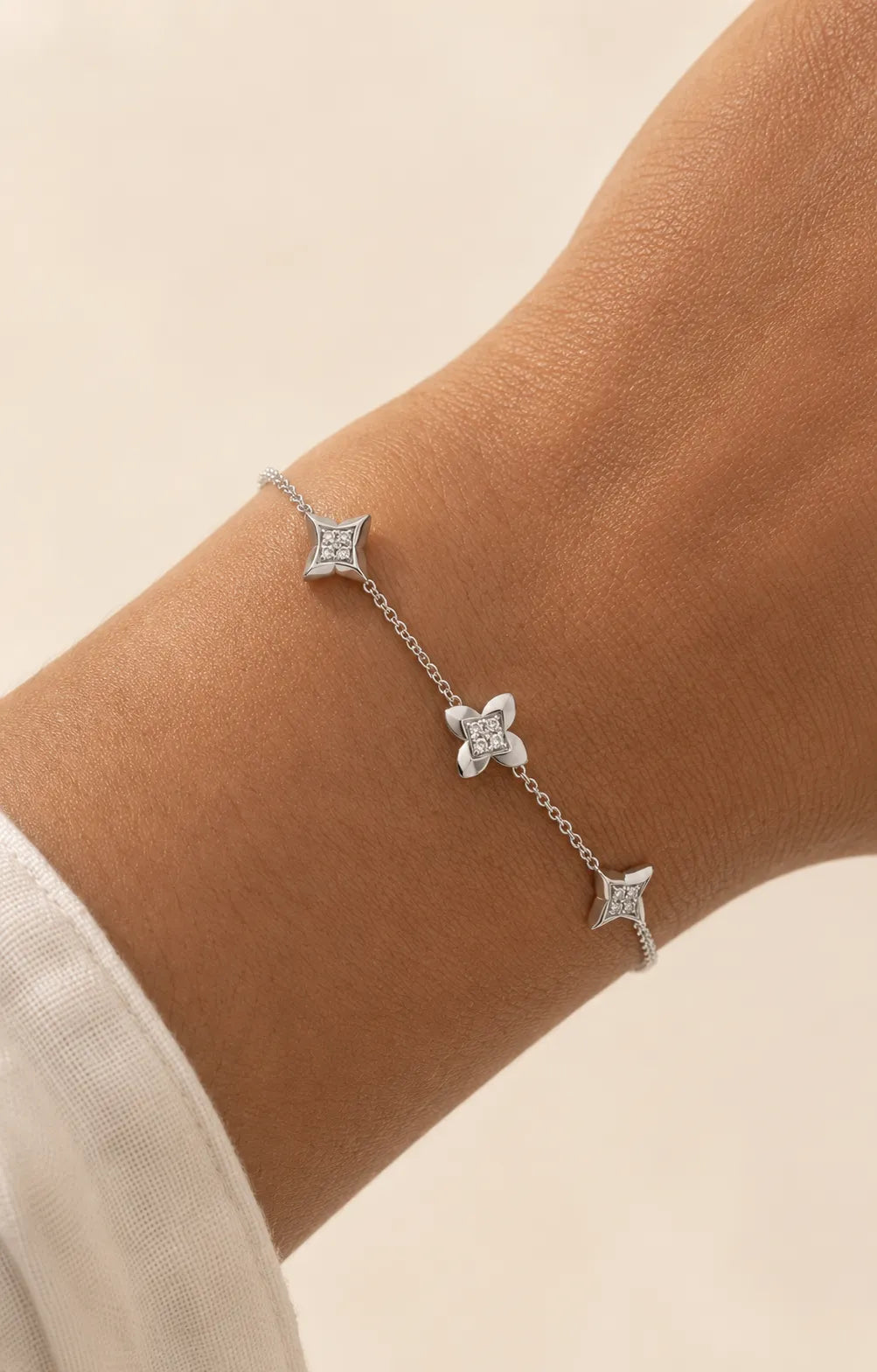 A delicate silver chain bracelet with three small star and flower motifs, shown worn on a person's wrist against a soft beige background.